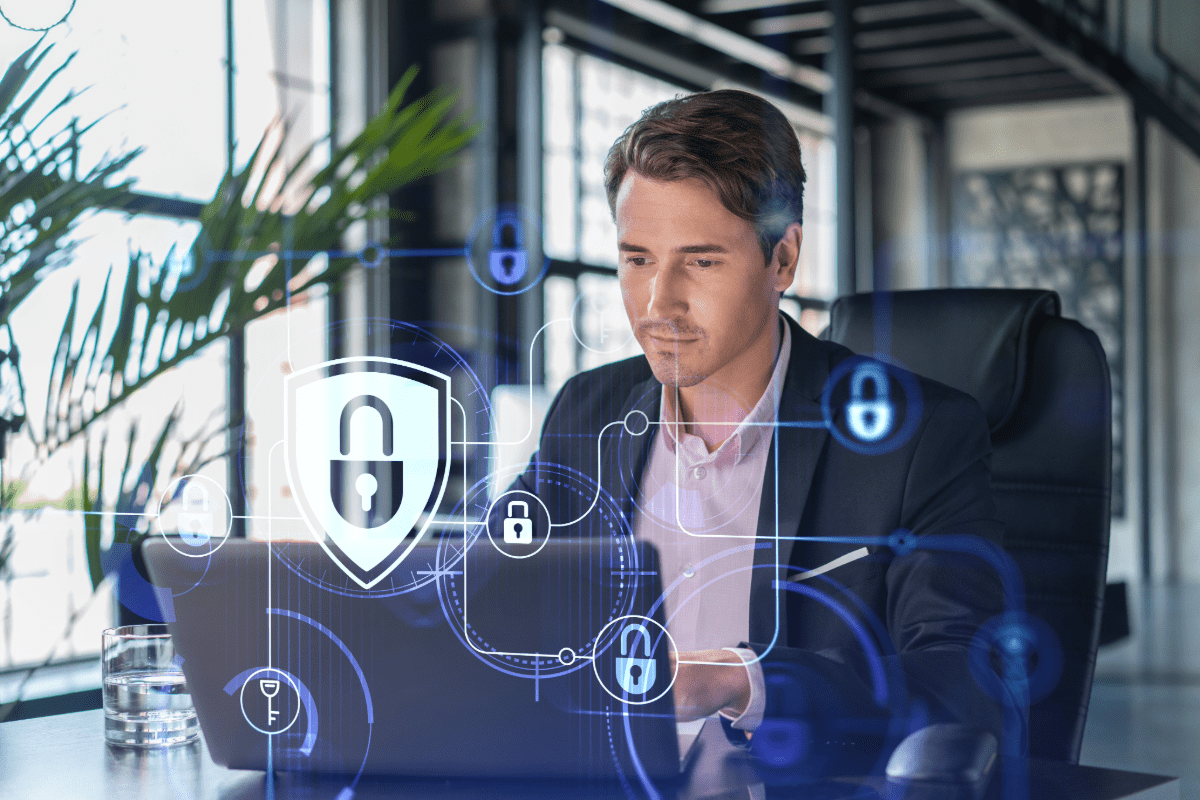 Businessman sitting at desk, looking at laptop computer screen, reviewing cyber liability insurance coverage. Lock and shield graphic overlay.