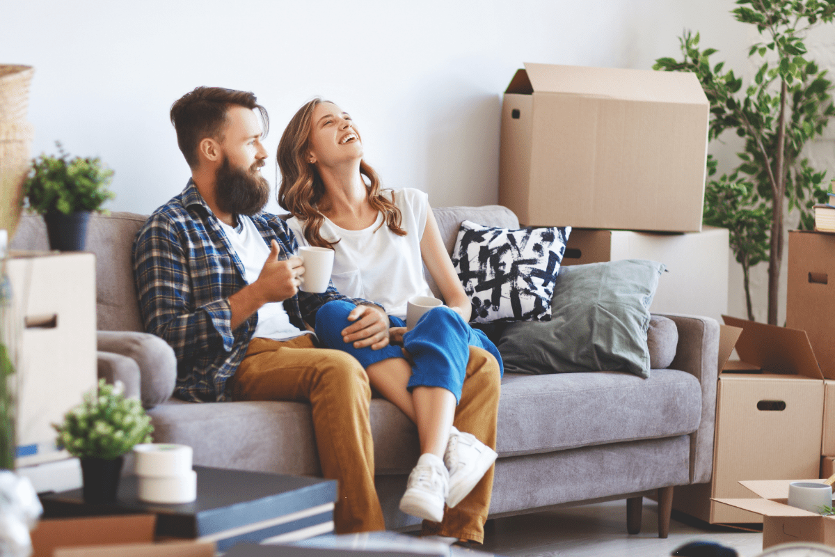A young couple sit on couch, coffee in hand, surrounded by moving boxes