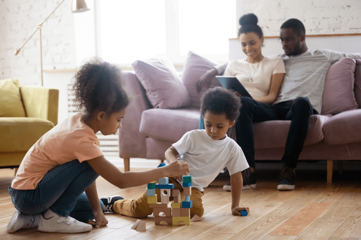 Two young siblings play with toys on the floor while thier parents research life insurance options on the couch behind them
