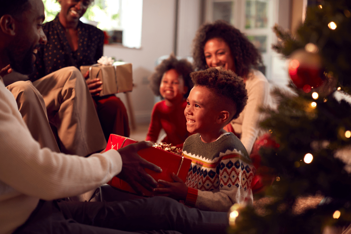 Adult handing child a present in front of a Christmas tree