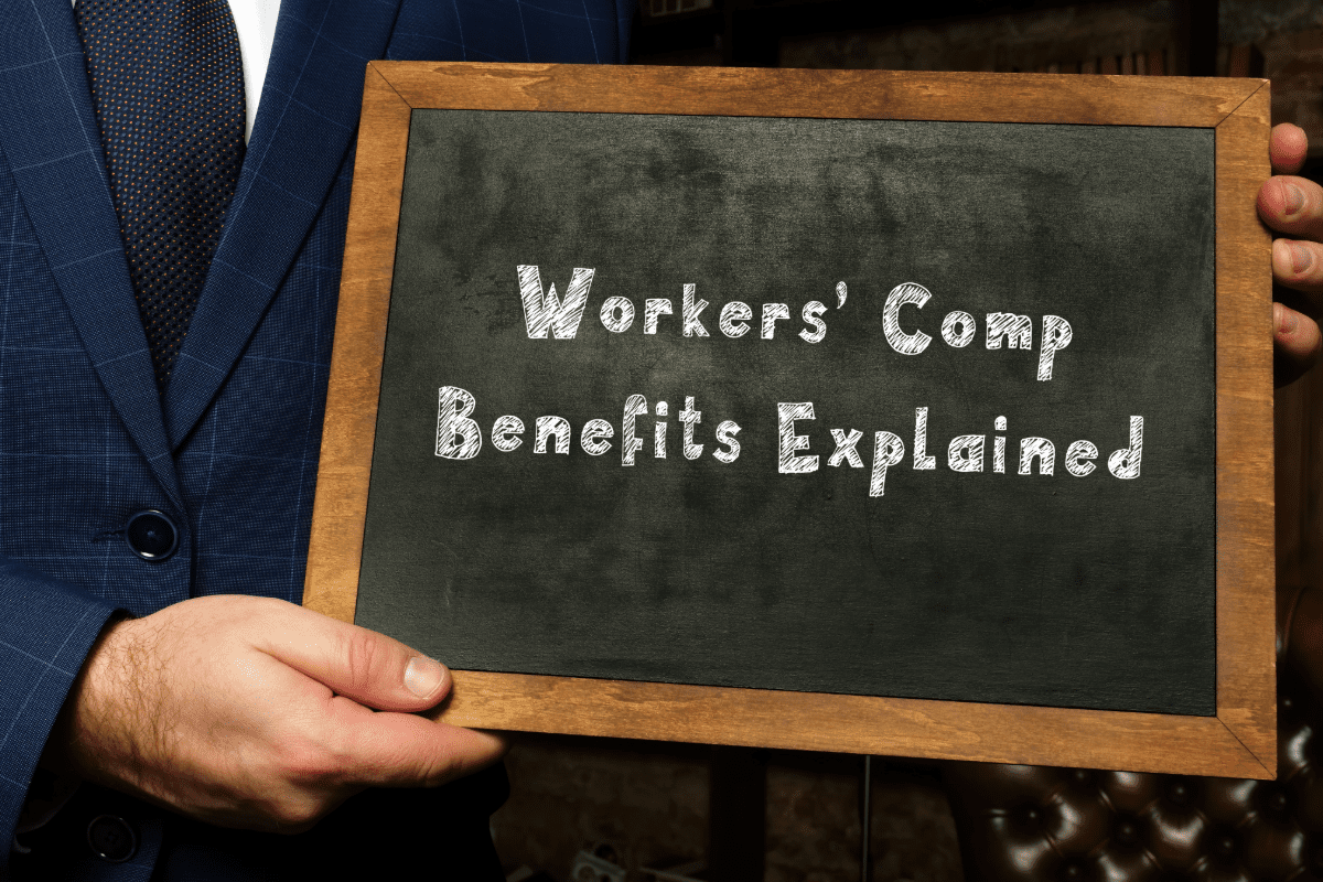 Hand holding chalkboard with "Workers' Comp Benefits Explained" written on it